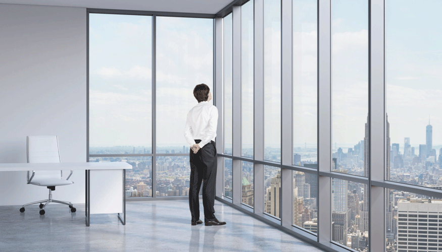 A man in a corner office room staring out floor-to-ceiling windows at a city